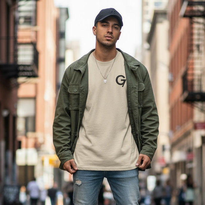 Man wearing a green jacket, beige t-shirt with a logo, and blue jeans on a city street.